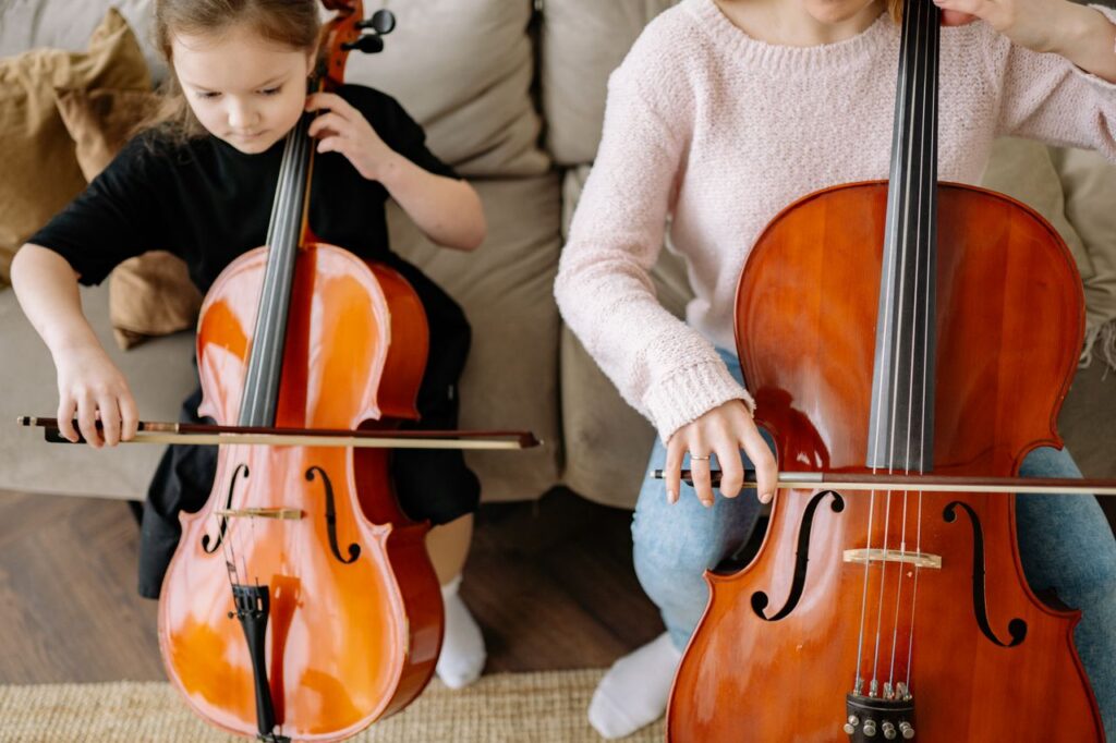 A young girl learning to play the cello alongside her mother, practicing music indoors.
