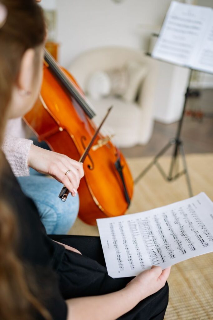Young girl learning to play the cello during a music lesson indoors, holding a music sheet.